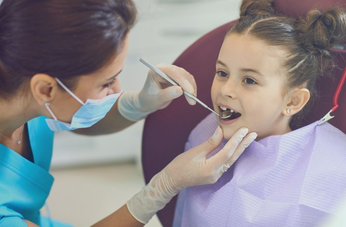 A dental hygienist wearing a blue uniform and face mask examines a young girl's teeth in a dental chair—highlighting the essential role dental industry recruiters play in staffing skilled professionals for patient care.