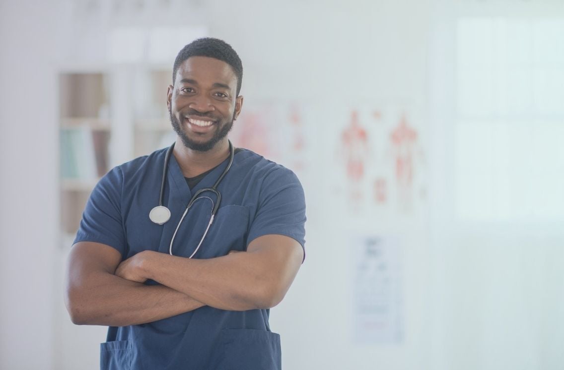 A smiling Licensed Practical Nurse (LPN) in navy blue scrubs with a stethoscope around his neck, standing confidently with arms crossed in a bright medical office 