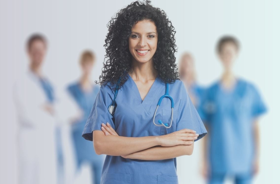 Smiling female nurse in blue scrubs with a stethoscope, representing the top talent nursing headhunters connect with leading healthcare organizations, with a blurred medical team in the background.